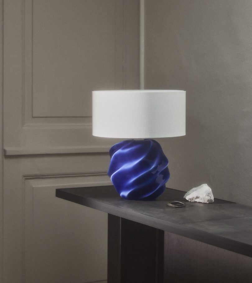 A table lamp with a white shade and a blue, sculpted base sits on a dark table next to a white rock and a pair of glasses in a minimalist room