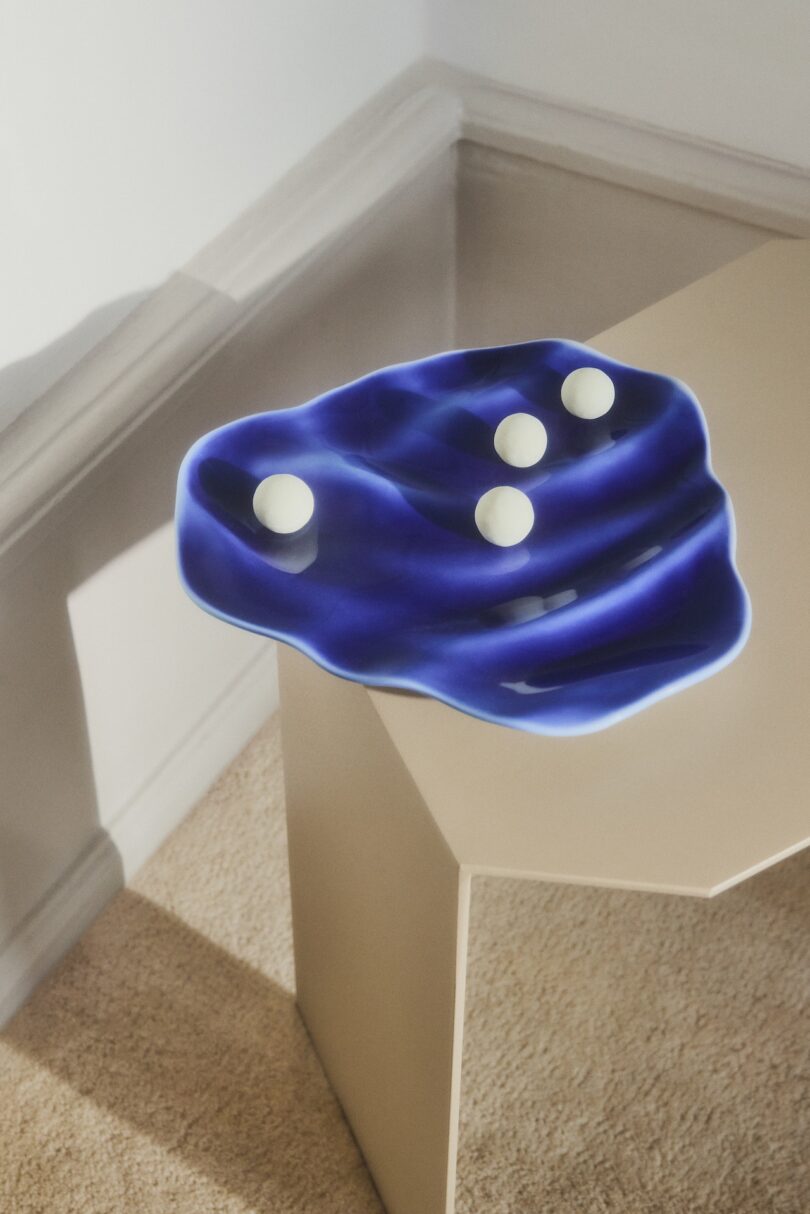 A blue, wavy, irregular-shaped tray with four white spheres rests on the edge of a beige table in a carpeted corner