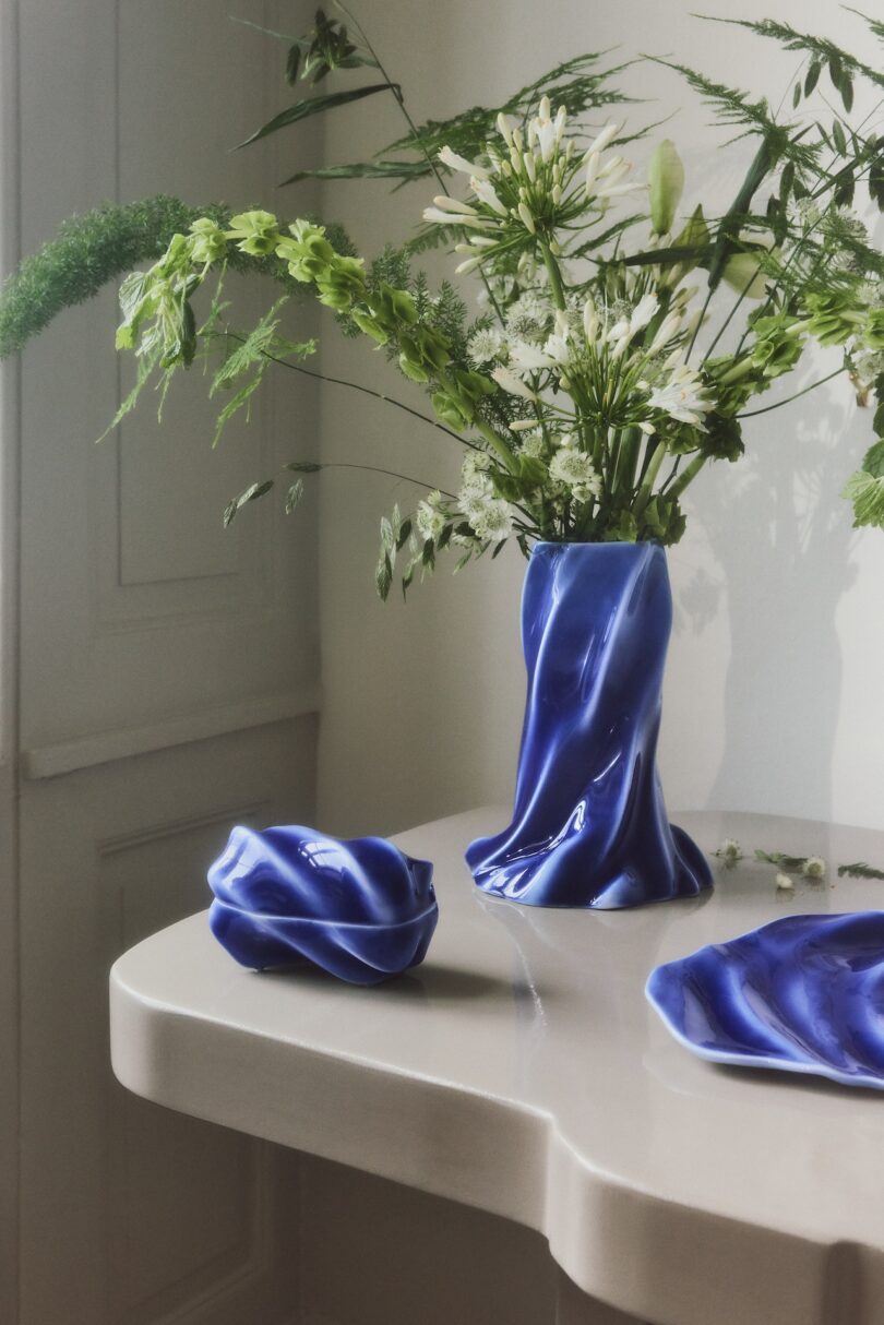 A blue, wavy ceramic vase with green and white flowers sits on a light-colored table next to matching blue ceramic dishes in a bright, minimal room