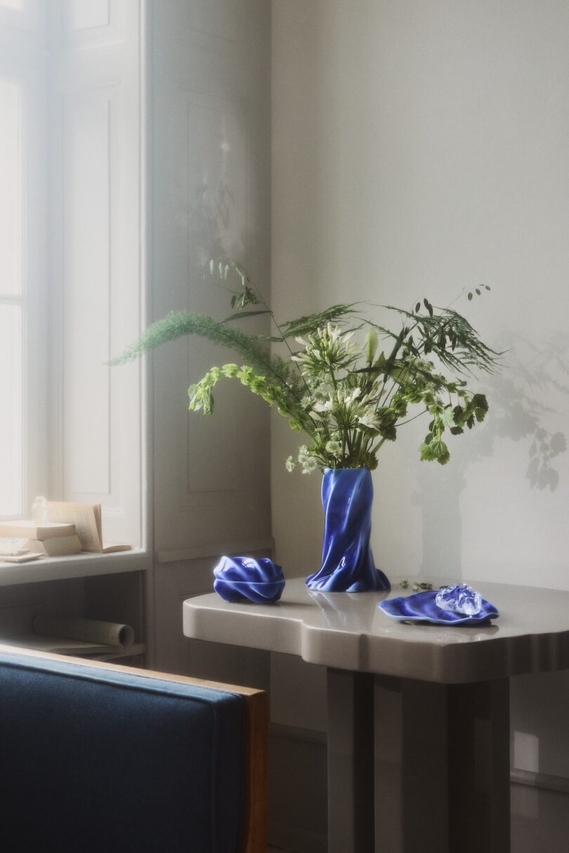 A modern table by a window holds a vase with green foliage and white flowers, a dish, and a dish with a lid