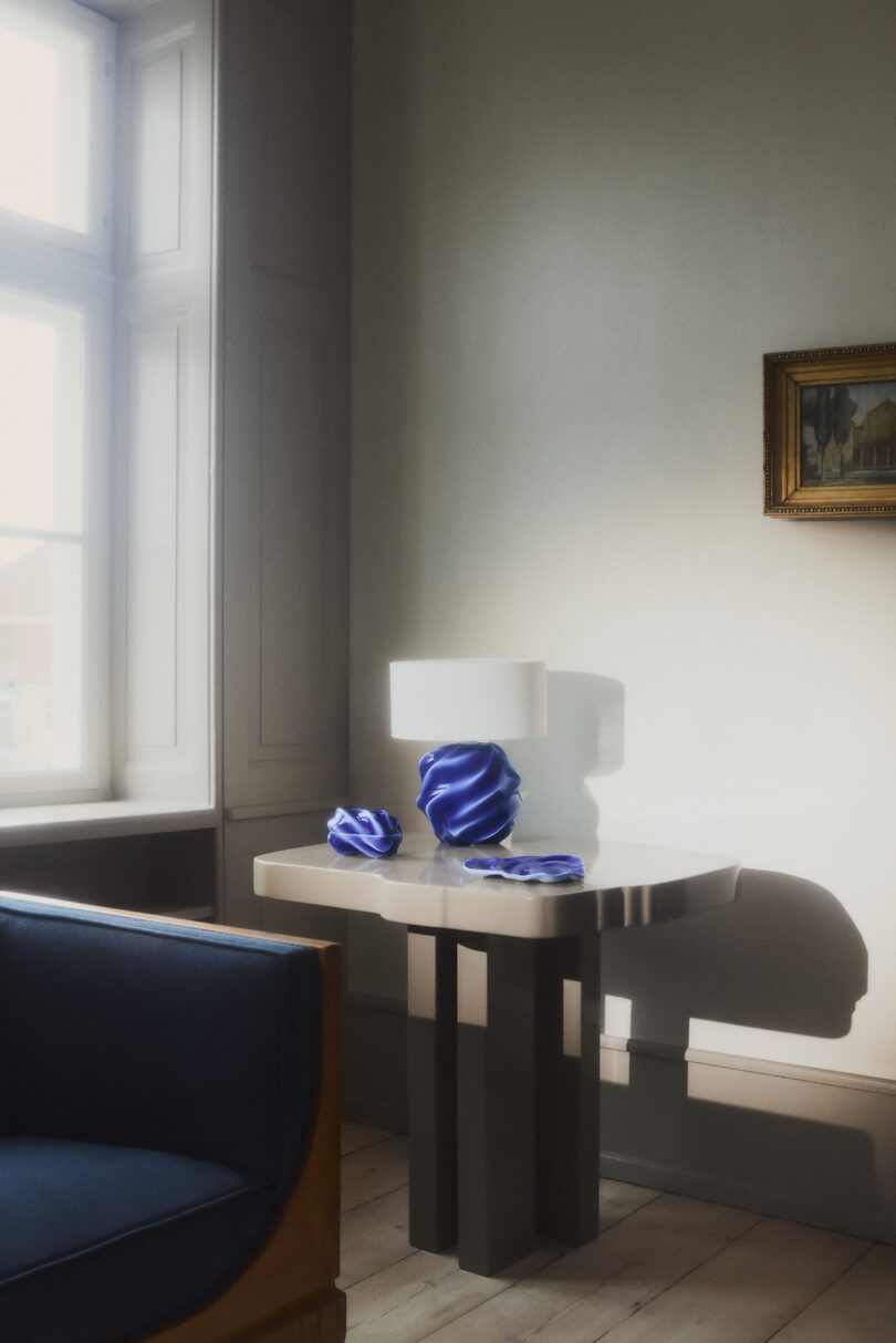 A modern table by a window holds a lamp , a dish, and a dish with a lid