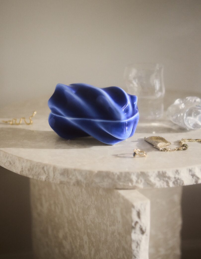 A blue sculptural object sits on a textured stone table alongside jewelry and two clear glass vessels