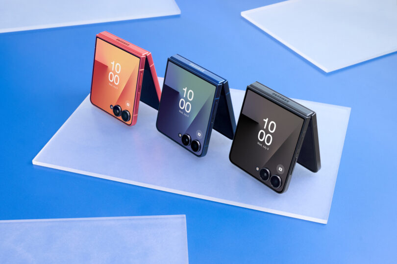 Three Samsung foldable smartphones in orange, blue, and black are displayed upright on a white surface against a blue background, each showing the time "10:00" on their exterior screens.