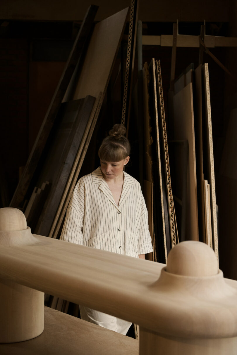 A person in a striped shirt stands in a woodshop, looking down at a large, unfinished wooden structure with panels and boards stacked in the background