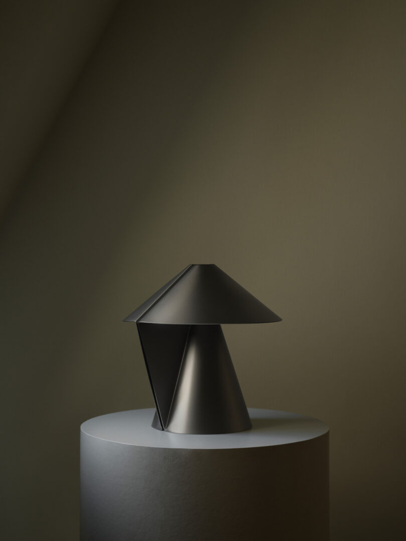 A modern, geometric black metal lamp sits on a round, white pedestal against a muted, minimalist background