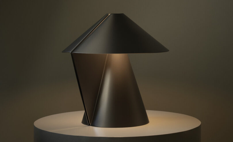 A modern black metal table lamp with a geometric, conical design sits on a round, light-colored surface, casting soft light downward