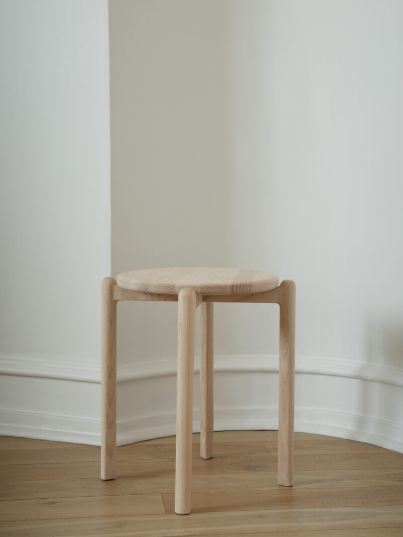 A light wooden stool with four legs is placed on a wooden floor against a white wall in a corner