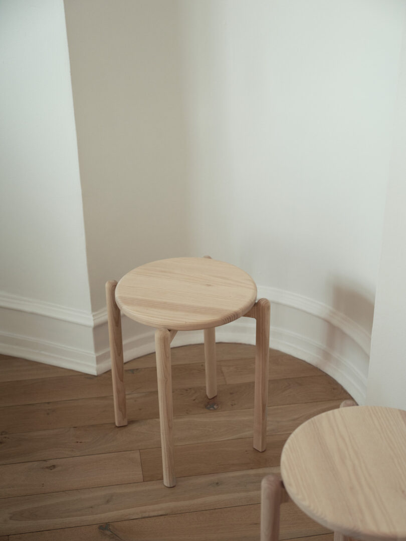A simple wooden stool with four legs is placed in the corner of a room with light-colored walls and wooden flooring. Another stool is partially visible in the foreground