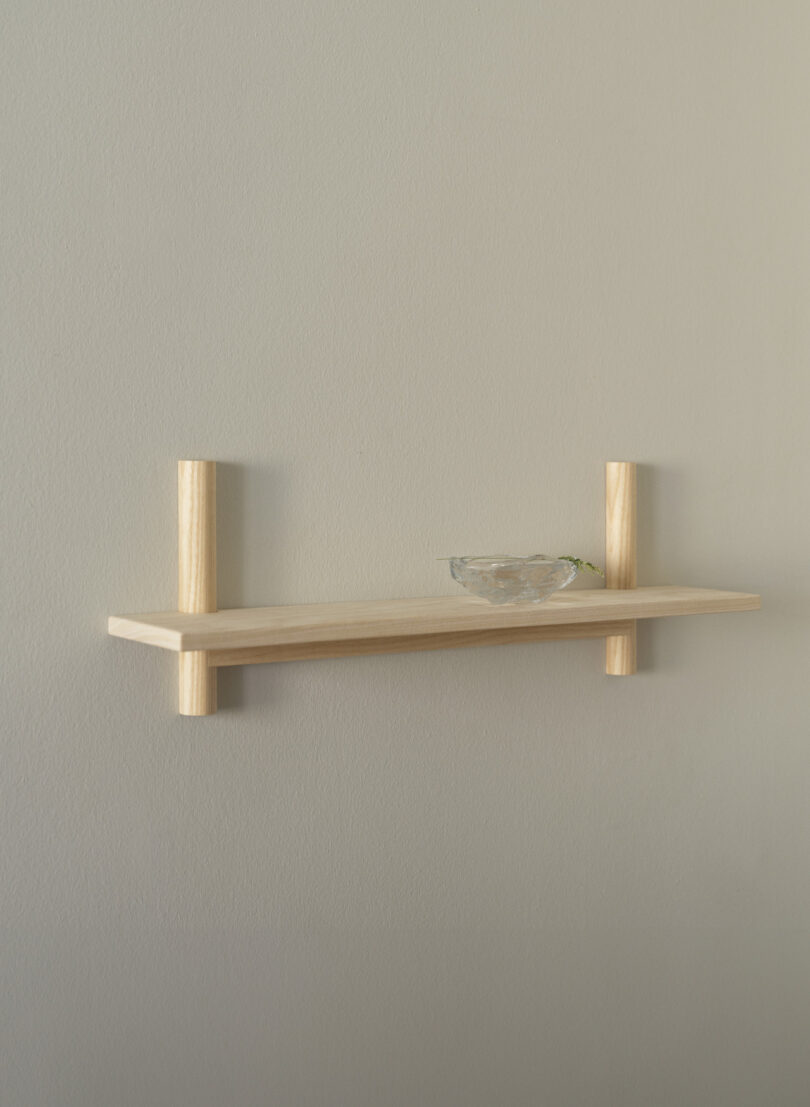 A light wood floating shelf is mounted on a plain beige wall, holding a small clear glass bowl