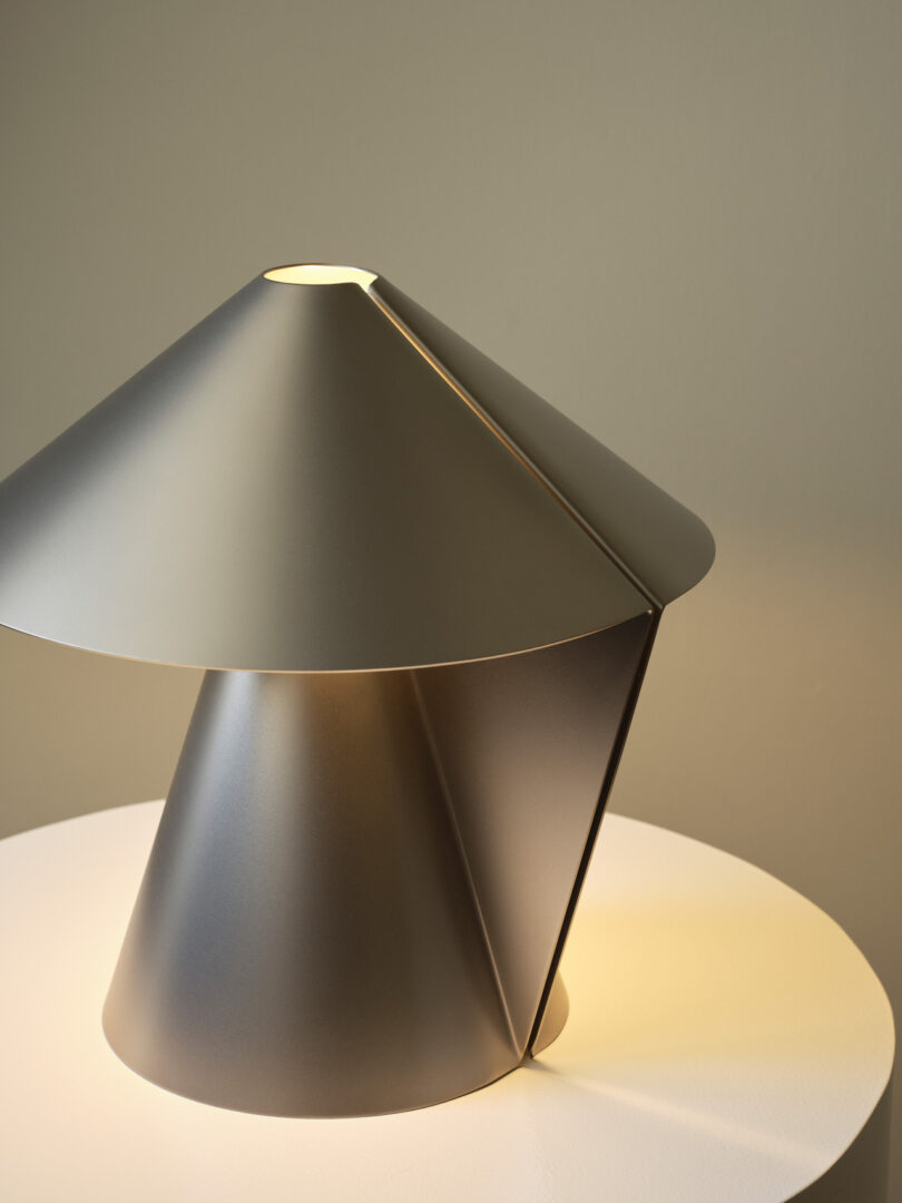 A modern table lamp with a metallic finish, featuring geometric shapes and a cone-shaped base, sits on a white surface with its light switched on