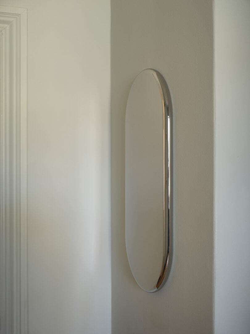 An elongated oval mirror with a metallic frame is mounted vertically on a light gray wall in a minimalistic interior space