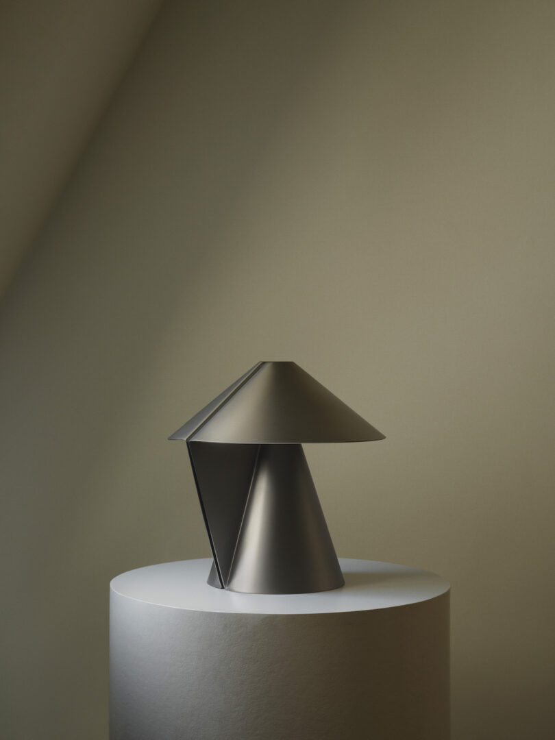 A modern, geometric black metal lamp sits on a round, white pedestal against a muted, minimalist background