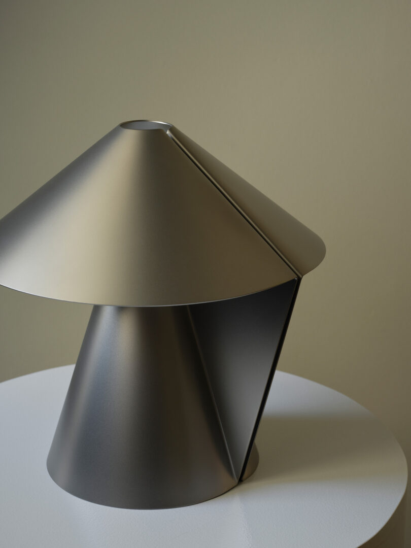 A metallic, modern table lamp with a conical shade and base sits on a round white surface against a neutral wall