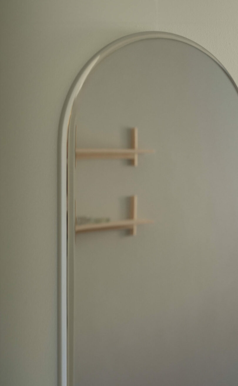 A close-up of a tall, arched mirror reflecting two minimal wooden wall shelves against a plain, light-colored wall