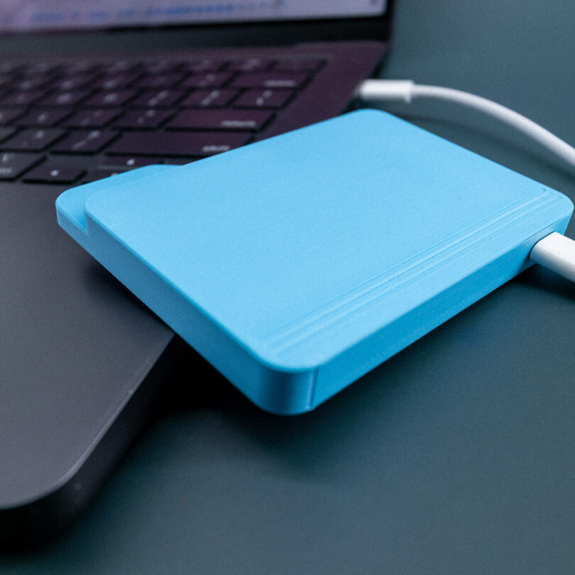 A blue external hard drive connected to a laptop via a white cable, placed on a dark surface.