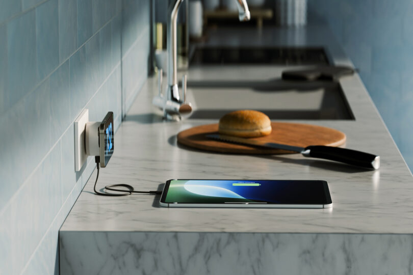 A tablet is charging on a marble kitchen counter near a cutting board with a bun and knife, powered by a TwelveSouth accessory. Nearby, a phone is plugged into a wall outlet.