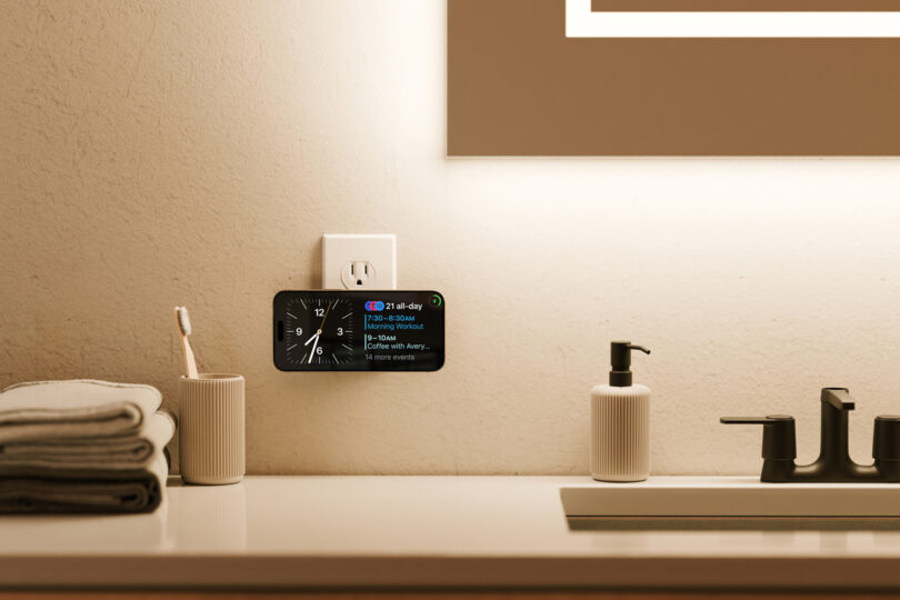 A bathroom countertop with folded towels, a toothbrush in a holder, a soap dispenser, and a smartphone mounted horizontally with a TwelveSouth accessory on the wall displaying the time and calendar.