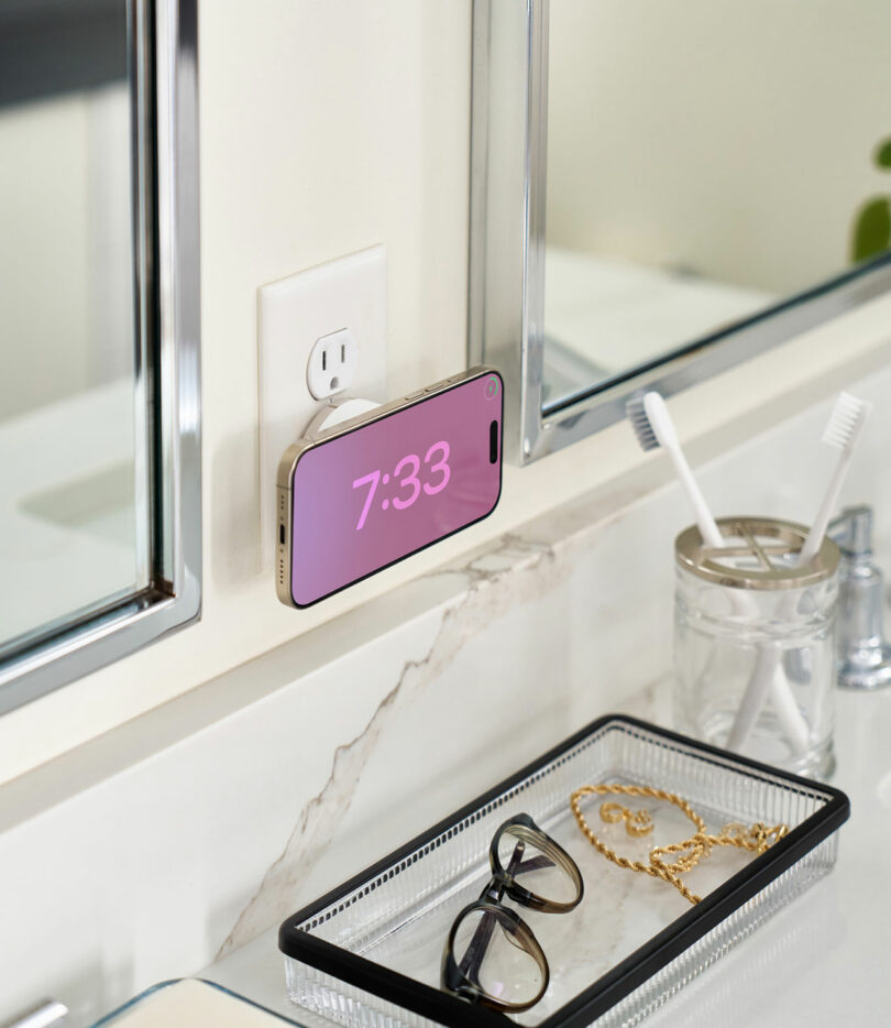 A TwelveSouth smartphone displays the time while plugged into a wall outlet above a bathroom counter with glasses, a toothbrush holder, and toiletries.