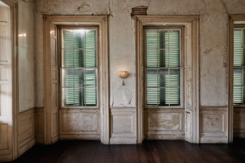 Aged interior wall with peeling paint, featuring two tall windows with green shutters, a wall sconce in between, and dark wooden floors.