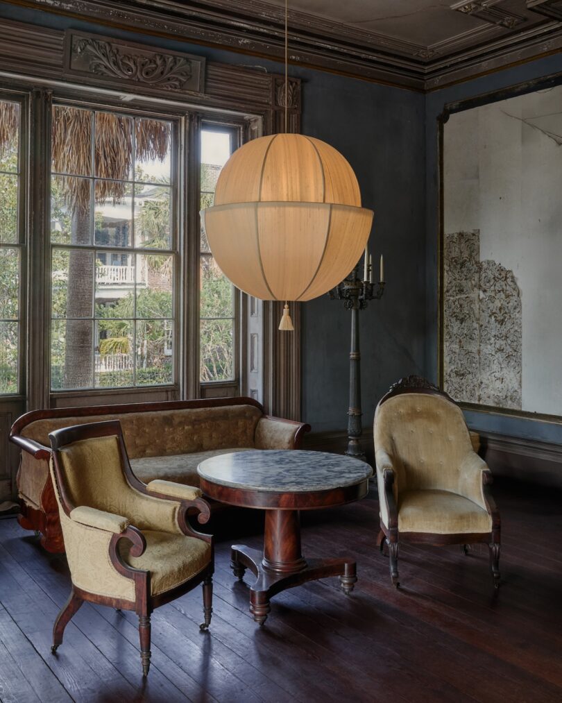 A vintage sitting room with antique chairs, a sofa, a round marble table, a large globe pendant light, tall windows, and a distressed mirror on the wall.