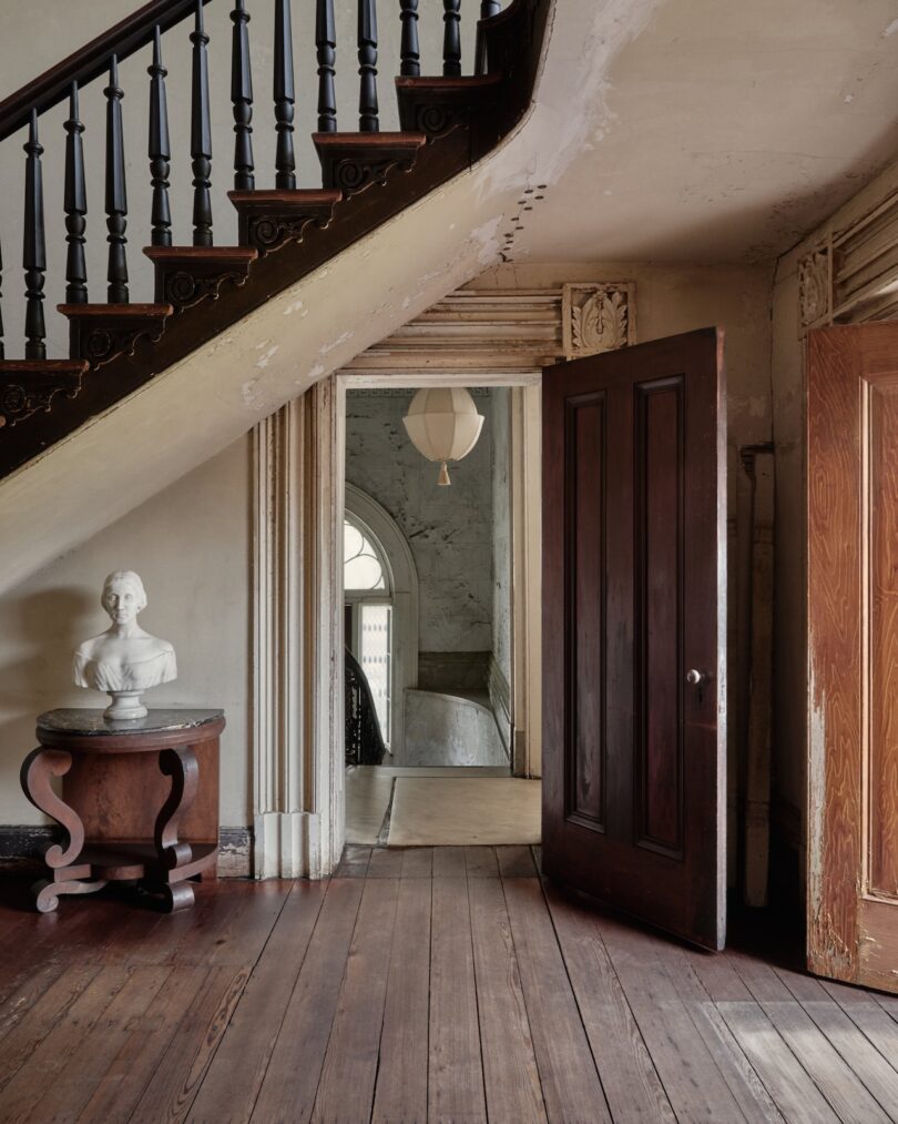 A wooden staircase with a dark banister rises above a small table holding a white bust. An open door reveals a hallway with arched windows and a hanging light fixture.