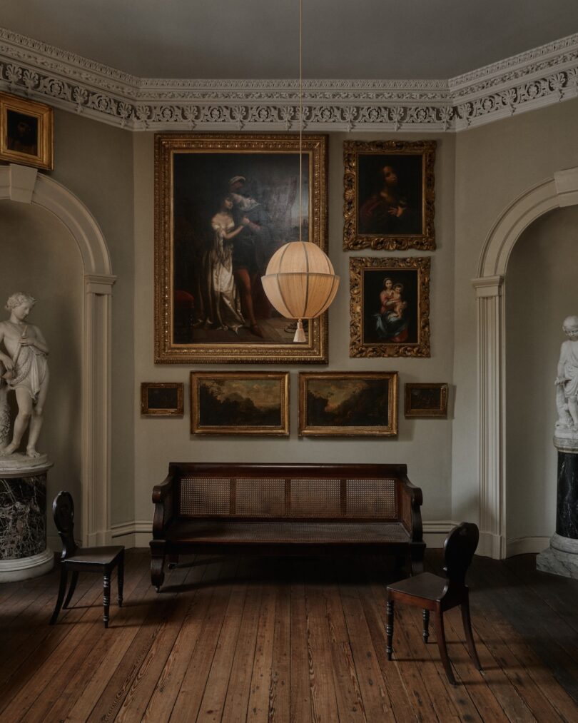 A classic room with a wooden floor, statues on either side, a central bench, two chairs, a hanging light, and several framed paintings on the wall.