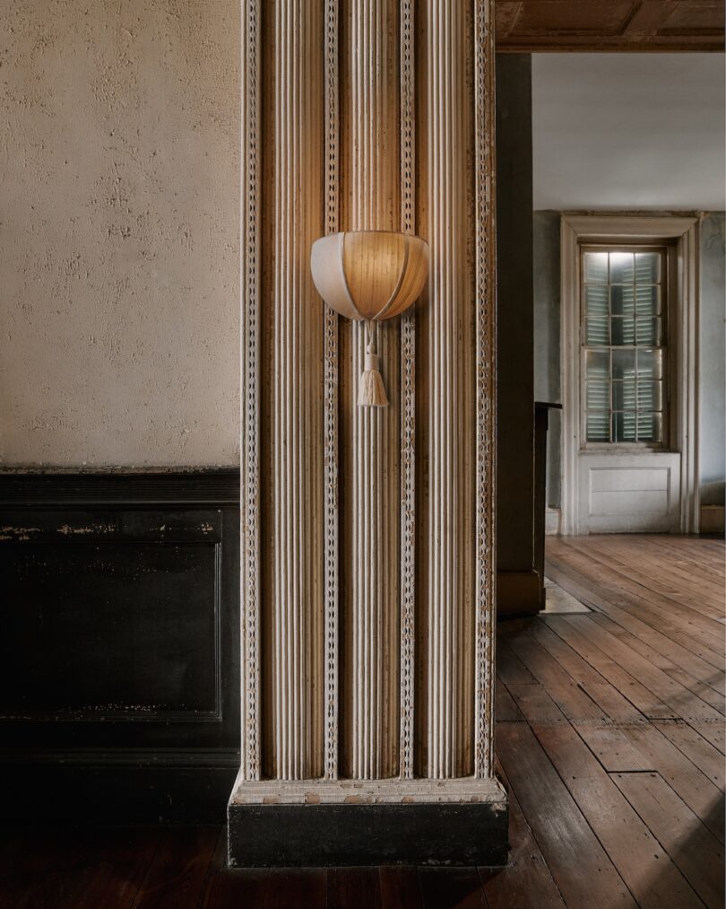 A sconce light fixture mounted on a fluted wall panel in a room with wooden floors, beige walls, and an open doorway leading to another space.