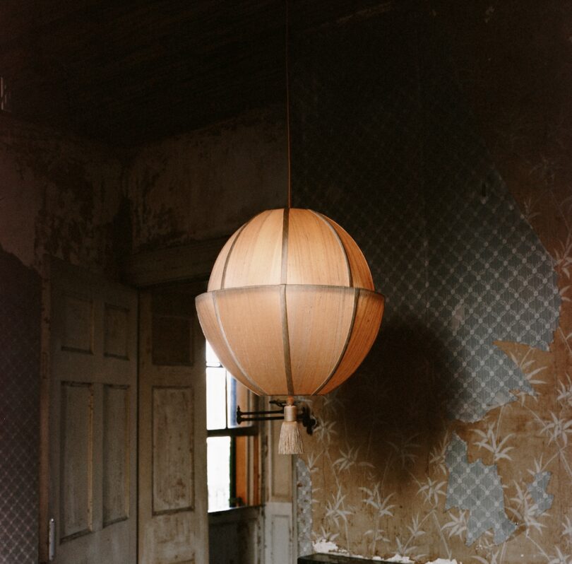 A spherical pendant light hangs from the ceiling in a dim, aged room with peeling wallpaper and a partially open door.