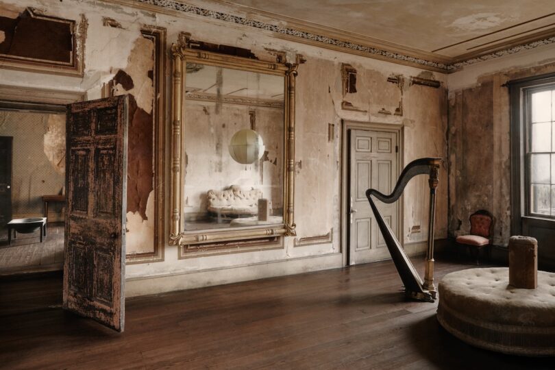 An old, worn room with peeling walls, a large ornate mirror, a harp, vintage furniture, and wooden floors under natural light from a window.