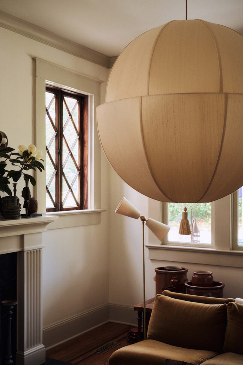 A large beige pendant light hangs in a cozy living room with a mustard sofa, floor lamp, fireplace, and wooden-framed windows.