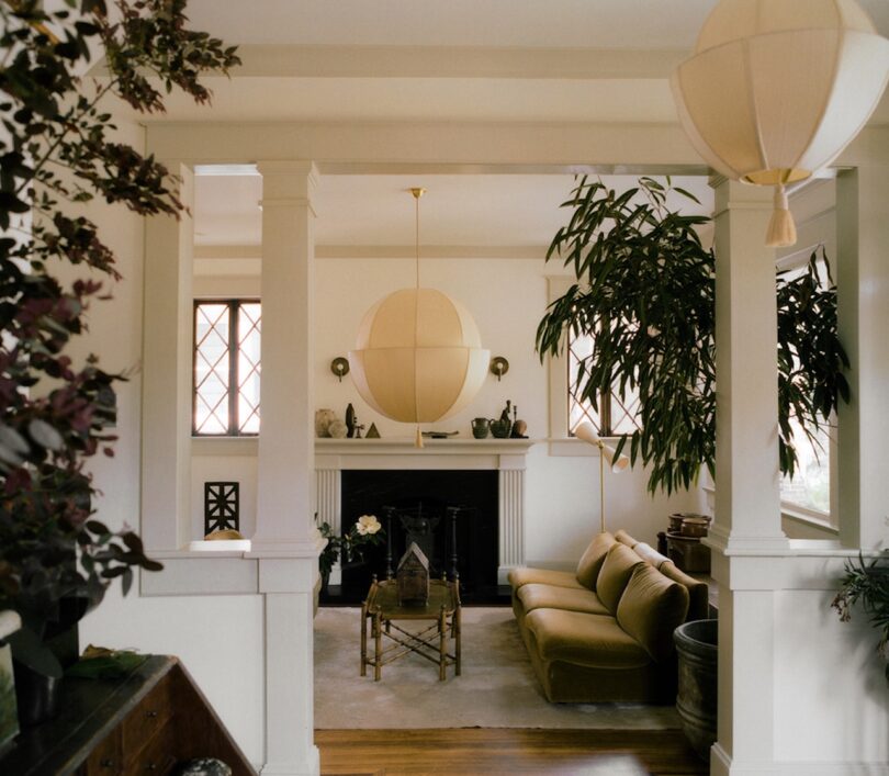 A living room with a beige couch, modern round chandelier, fireplace, large potted plants, and windows letting in natural light.