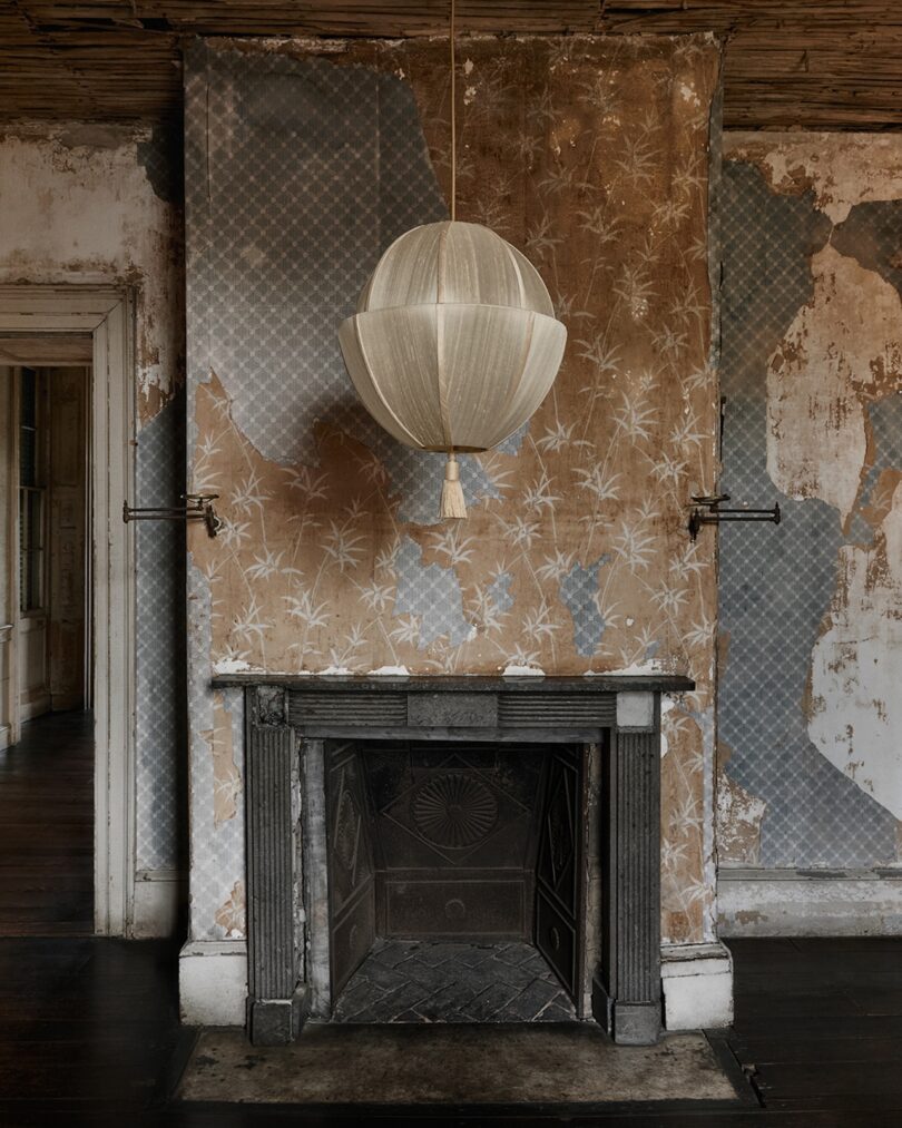 A vintage fireplace with a decorative mantel sits below a modern fabric pendant light; the walls have peeling, patterned wallpaper, showing signs of age and wear.