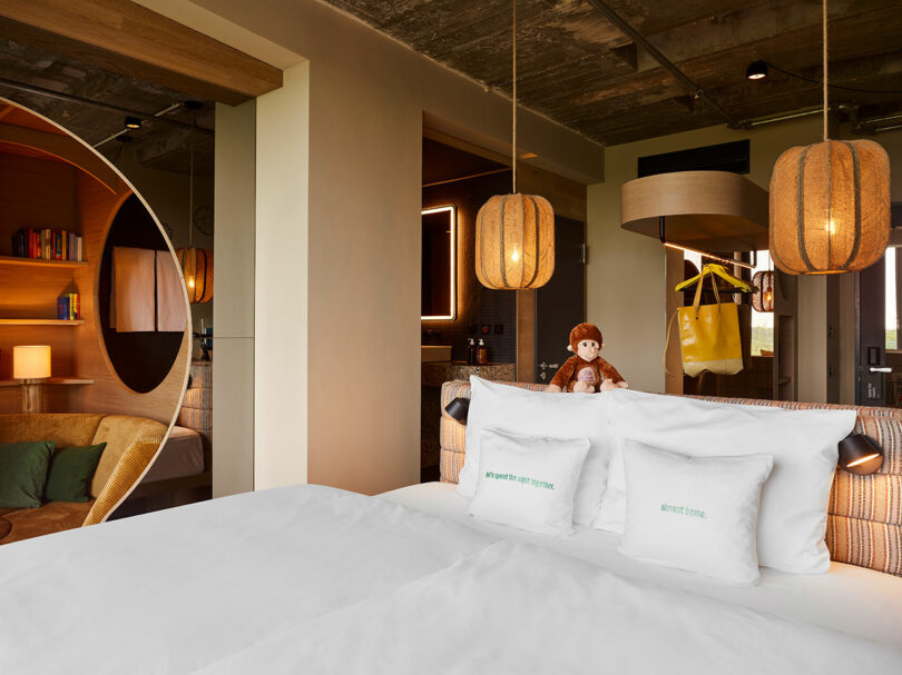 A modern 25hours hotel room with a large bed, two white pillows, a plush monkey toy, pendant lights, and a cozy reading nook featuring shelves and a round opening.