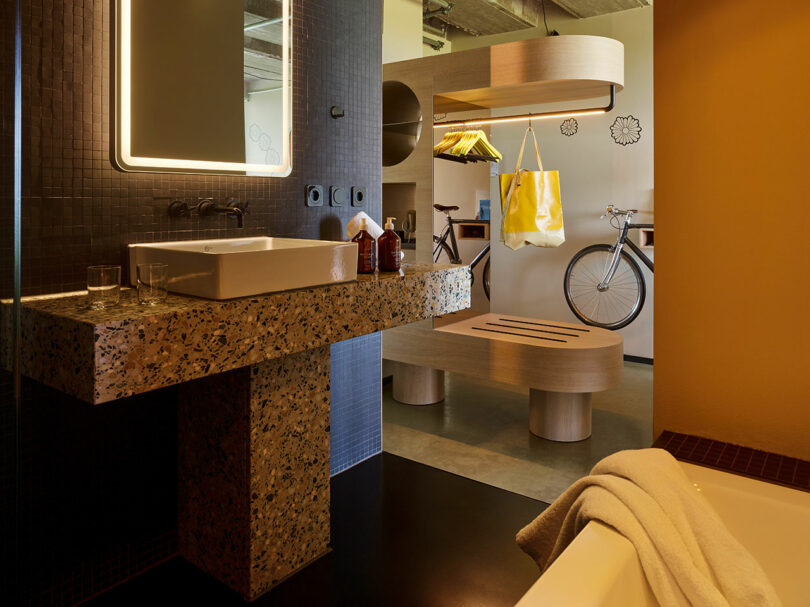 Modern 25hours bathroom with a terrazzo sink, illuminated mirror, and a view into a room featuring a bench, hanging clothes, shelves, and a bicycle against the wall.