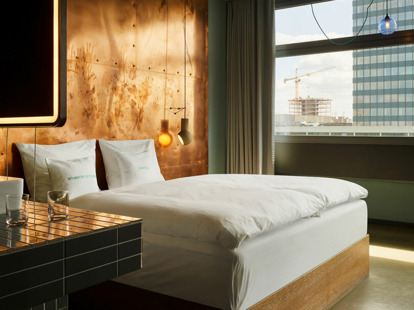 Modern 25hours hotel room with a large bed, copper accent wall, hanging lights, glass of water on a tiled counter, and a window revealing city buildings and a crane outside.