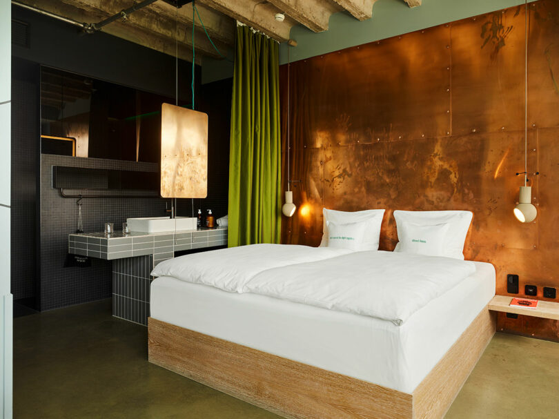 A modern 25hours hotel room with a double bed, white bedding, a copper accent wall, green curtain, hanging lights, and a tiled sink area.