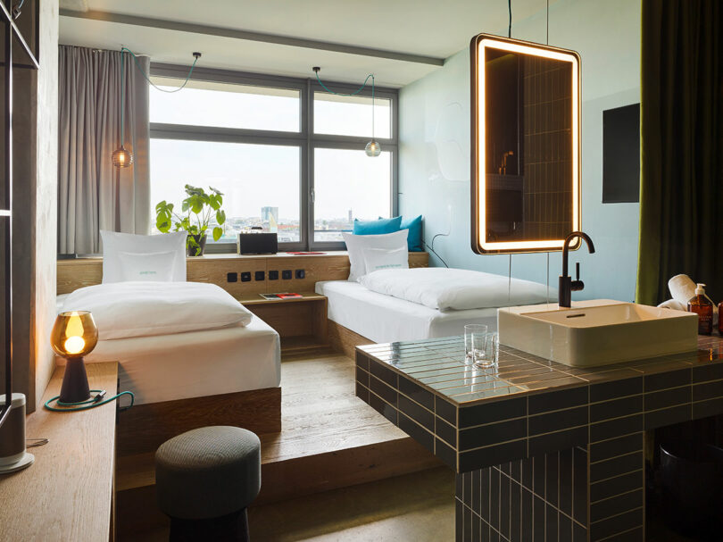 Modern 25hours hotel room with two single beds, a large window, plants, a glowing lamp, bedside table, and a tiled bathroom vanity featuring an illuminated mirror and sink.