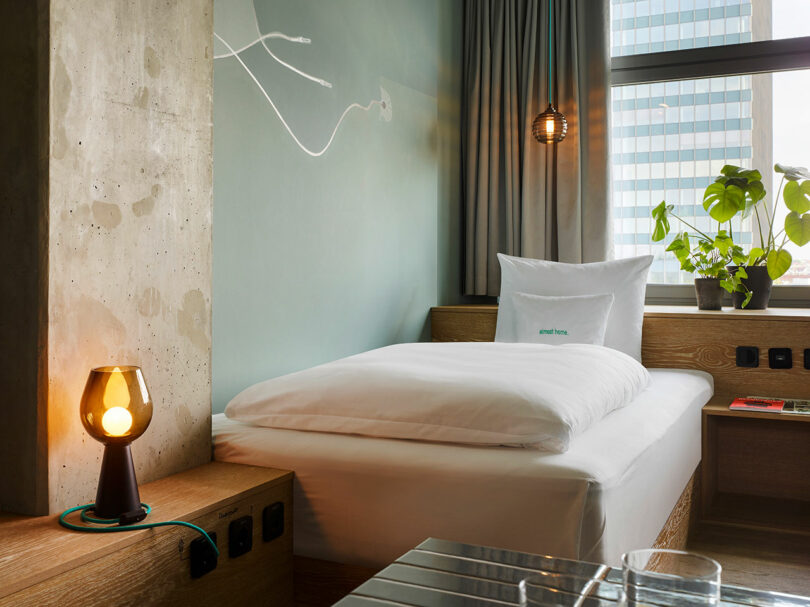 Modern 25hours hotel room with a single bed, potted plant, bedside lamp, and large window showing a cityscape. Neutral wall colors and minimalistic decor create a relaxed atmosphere.