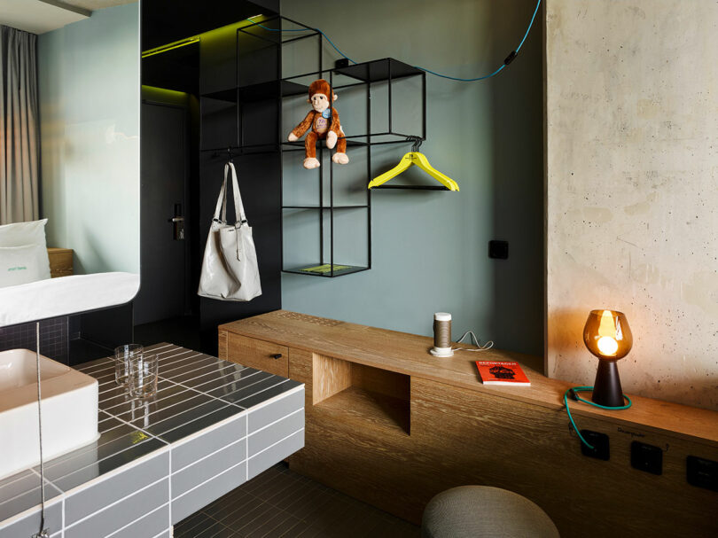 A modern 25hours room with minimalist shelves holding a white bag, a hanging yellow hanger, a toy monkey, a book, a smart speaker, and a lit lamp on a wooden surface.