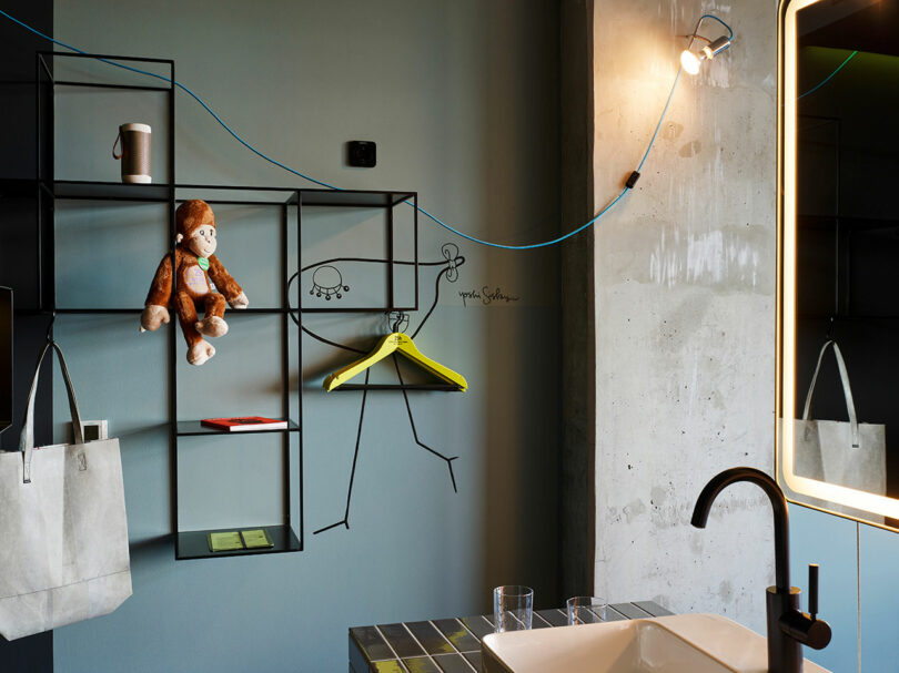 A modern bathroom inspired by 25hours features a black faucet, a grid wall shelf with a stuffed monkey, a cup, a yellow hanger, and a wall doodle, plus a light attached by a blue cord.