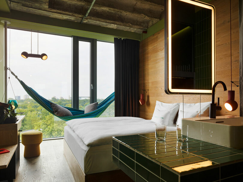 A modern 25hours hotel room with a large bed, a wall-mounted sink, a green-tiled countertop, and a hammock by the window offers relaxing views of trees outside.