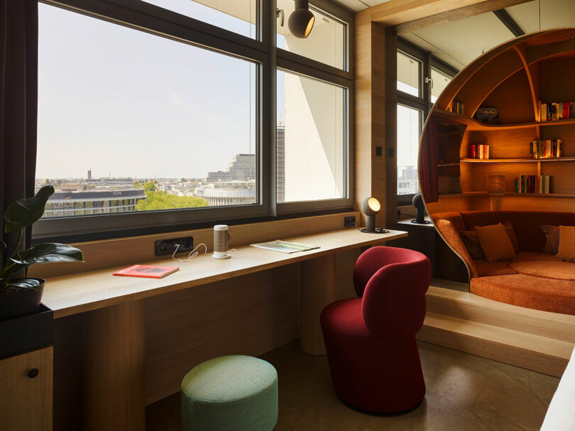 A modern 25hours workspace with a long wooden desk, two chairs, a plant, and a built-in circular bookshelf seating area by large windows overlooking a cityscape.