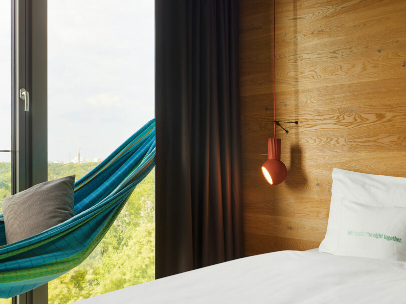 A bright 25hours room with wooden walls features a white bed, a blue hammock by the window, a pillow, dark curtains, and a red hanging lamp.