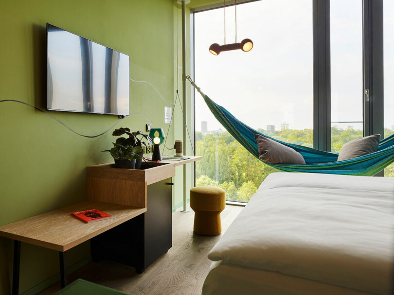Modern 25hours hotel room with green walls, a bed, wall-mounted TV, and a hammock by large windows overlooking trees. Features include a small desk with plants and a bench with a book.