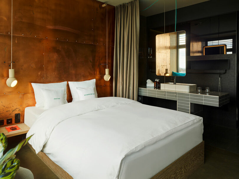 Modern 25hours hotel room with a double bed, copper accent wall, hanging lights, and an open bathroom area featuring a sink and mirror.