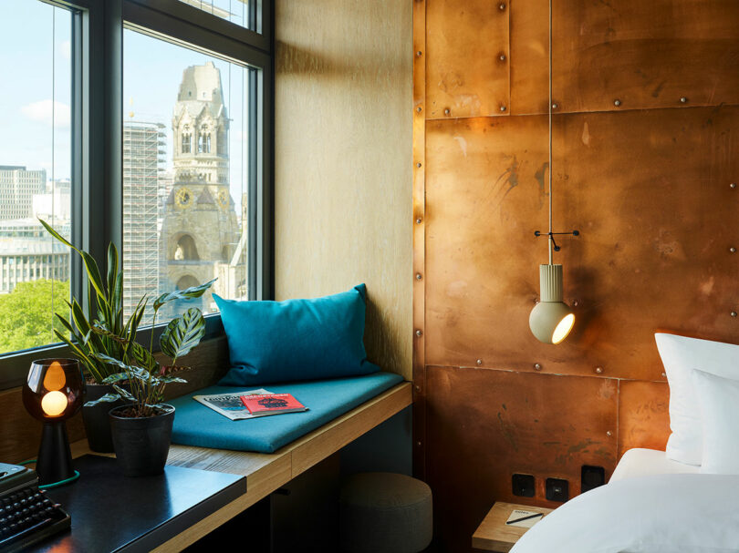 A modern 25hours hotel room featuring a copper accent wall, a bed, a desk with a plant and lamp, plus a window seat overlooking the cityscape and its historic church tower.