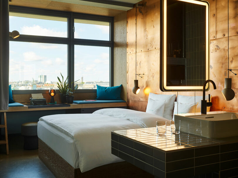 A modern 25hours hotel room with a large bed, built-in desk by a window, illuminated mirror, sink, and cityscape view outside.