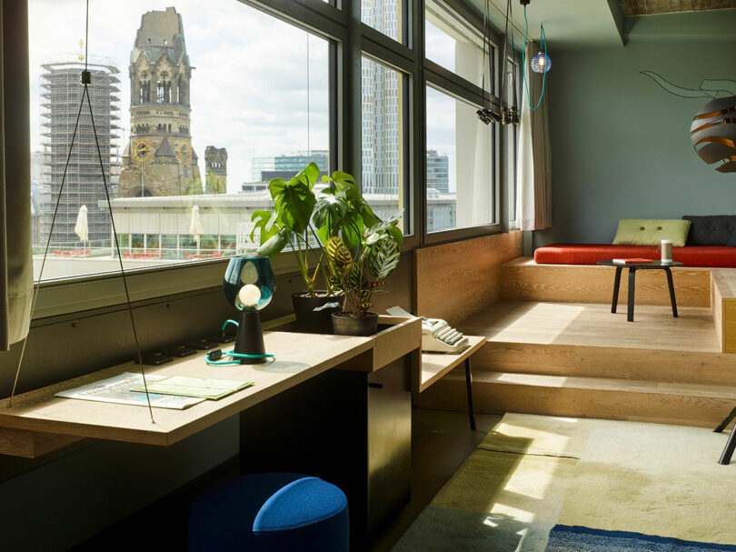 A bright modern 25hours room with a desk, houseplants, a lamp, and large windows overlooking city buildings, including a historic church tower in the background.