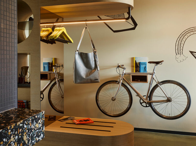 A modern 25hours interior features a bicycle mounted on the wall, a large metallic bag hanging, open shelving with books, and clothes hangers under a wooden shelf with integrated lighting.
