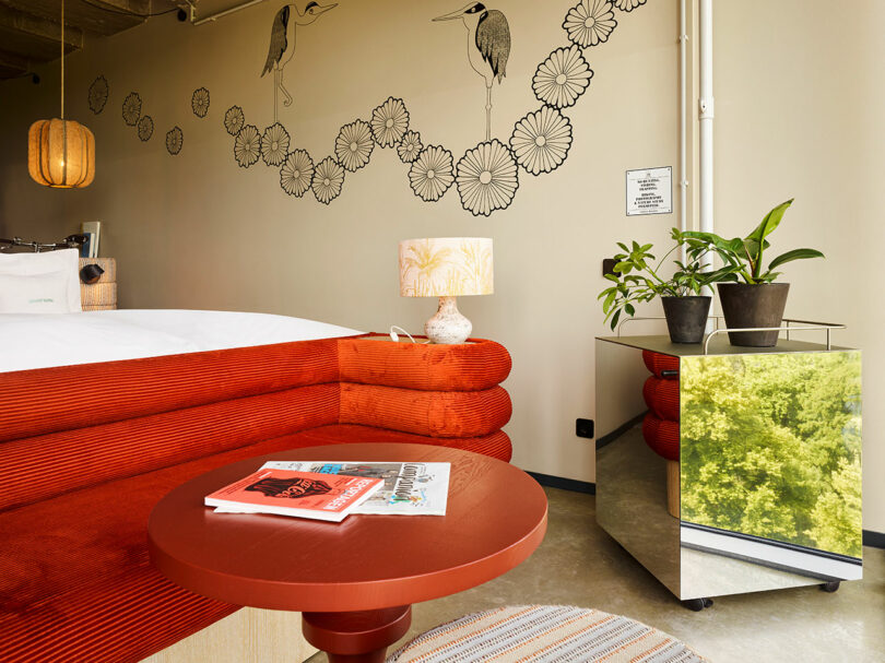 Modern 25hours hotel room with a red velvet sofa, round table with magazines, potted plants on a mirrored side table, and wall art featuring birds and flowers.